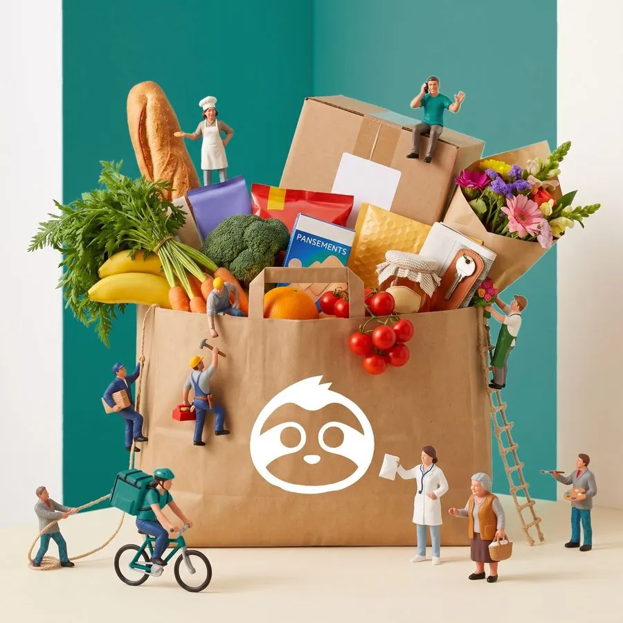 Un grand sac Allô La Flem rempli de pain, de fruits, de légumes, de fleurs et d'un colis. Autour, des figurines miniatures : une boulangère, des artisans, un fleuriste, une pharmacienne, un peintre, un concierge au téléphone et une livreuse à vélo, au service d'une dame âgée.